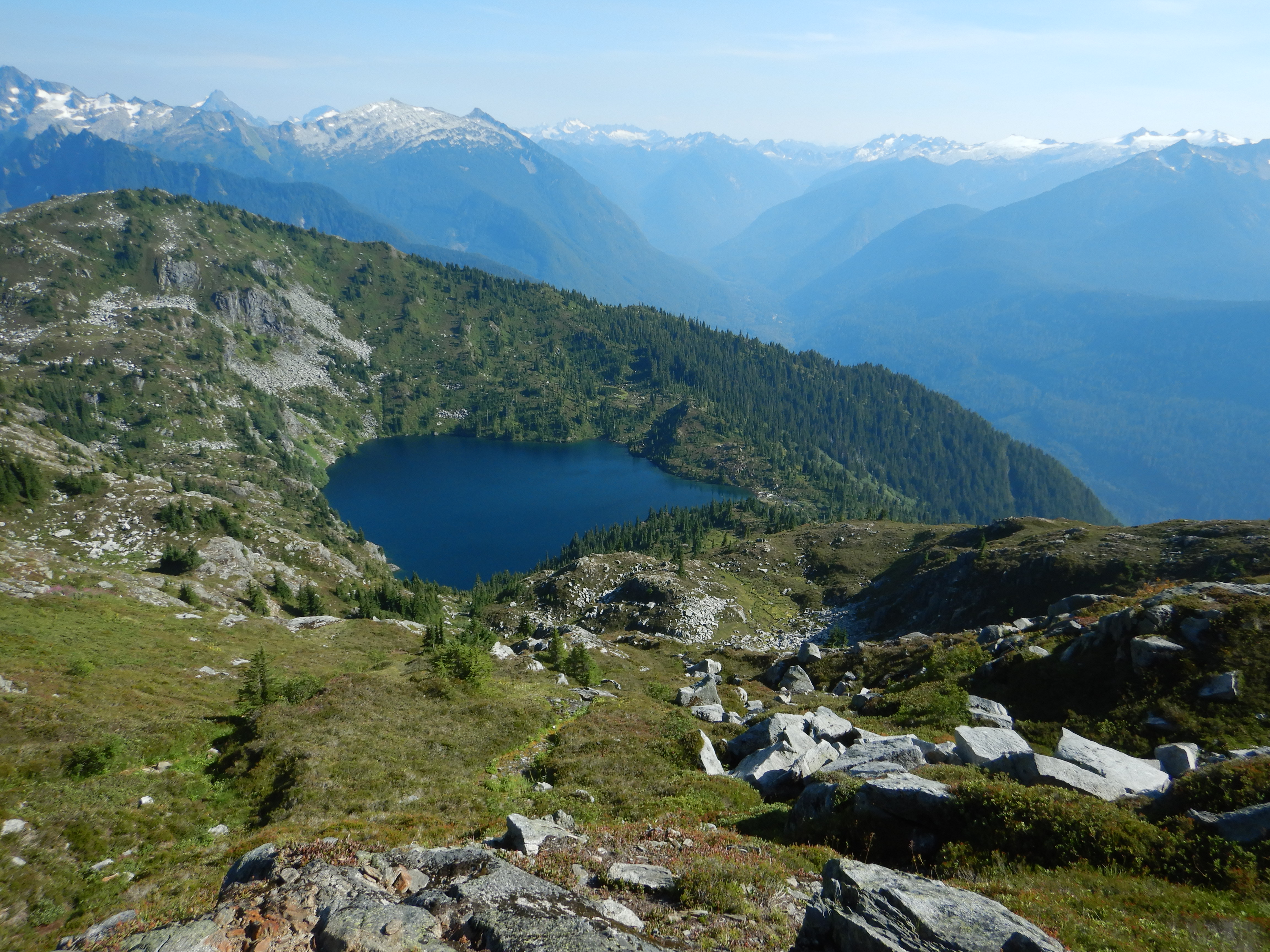 View looking toward a lake in a glacial cirque. Deep valley and snow covered peaks on horizon.