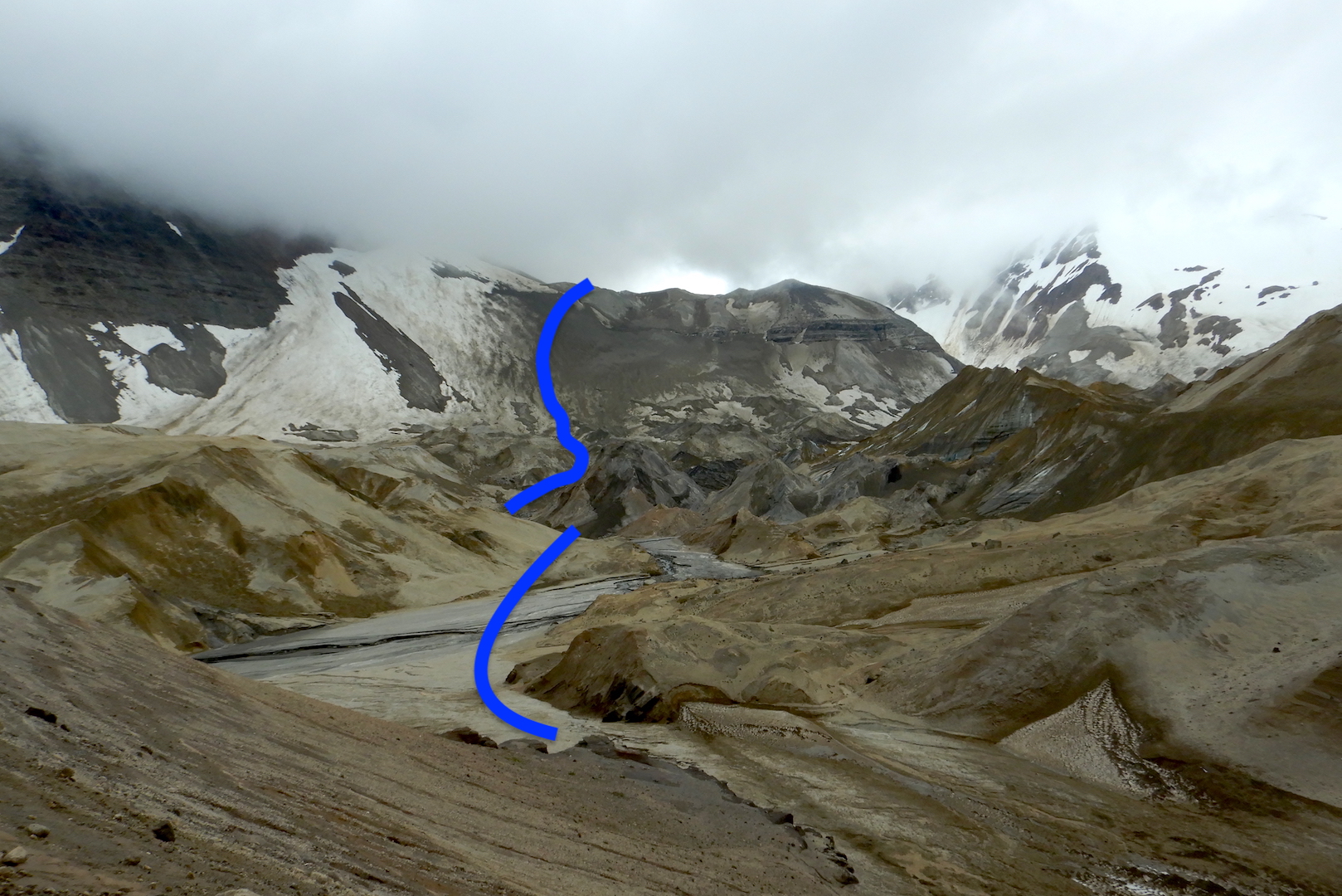 View of hummocky landscape created by ash and pumice covered glaciers at the foot of mountains hidden in clouds. Blue line near center represents route.