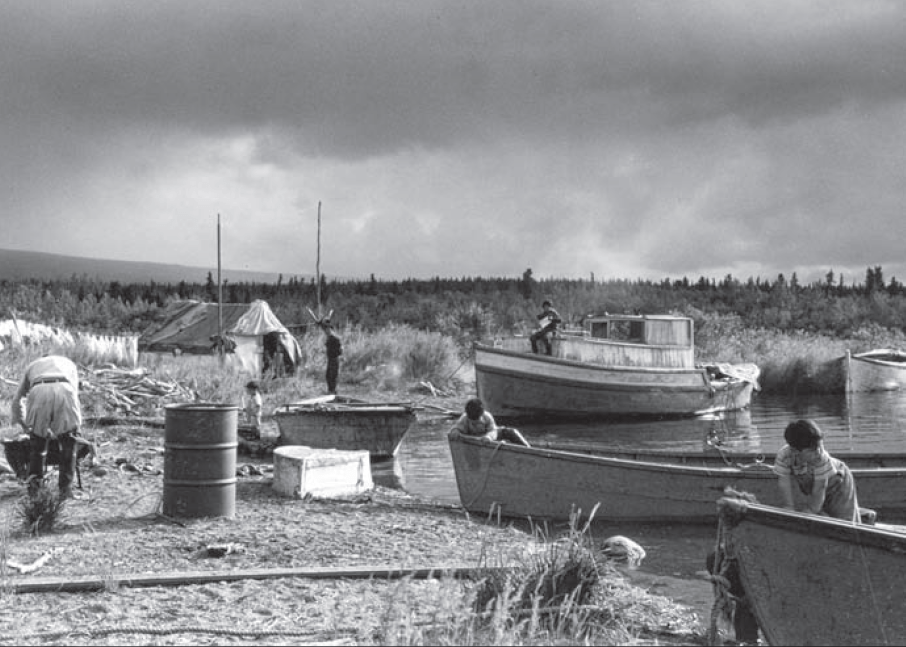black and white photo of Alaska Native families on a beach. Four boats are in the water at right. Children and adults stand on and near the boats.