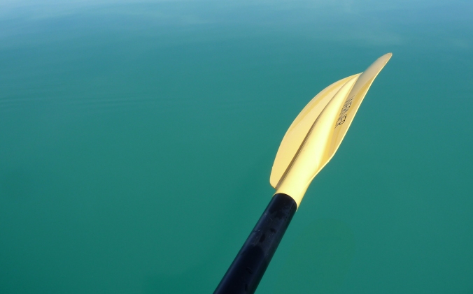 turquois colored lake water. A yellow kayak paddle with a black handle offers contrast