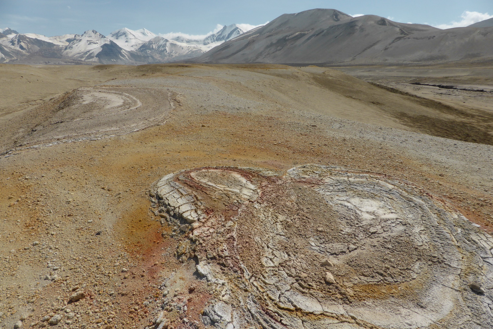 ovoid crusts of clay (lower right) lay on top of gray, orange, and brown pumice fields. snow covered mountains