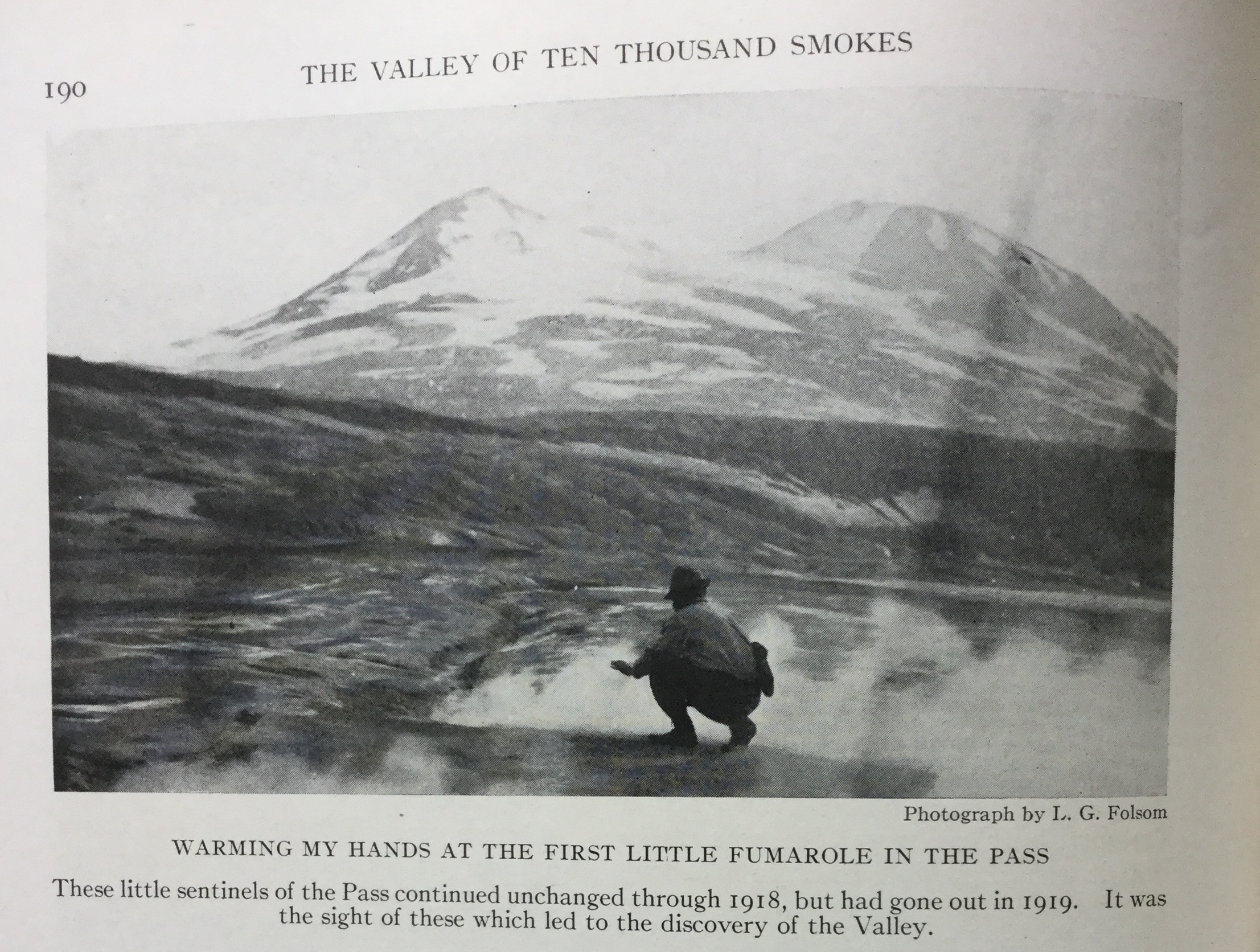 black and white photo. man squats at center next to small steam vent. a snow capped mountain is o the horizon. text reads "190. The Valley of Ten Thousand Smokes. Photograph by L. G. Folsom. Warming my hands at the first little fumarole in the pass. These little sentinels of the Pass continued unchanged through 1918, but had gone out in 1919. It was the sight of these which led to the discovery of the Valley."