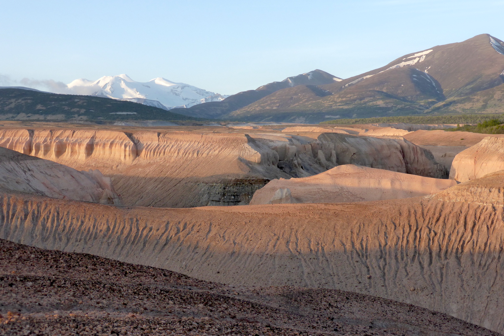 canyons carved through pumice and ash on lower half of photo. mountains line horizon. low angled sunlight highlights depth of canyons