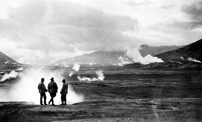 three men stand in front of fumarole at lower left. A valley filled with many steaming fumaroles fills the rest of the middle ground. clouds and mountains fill the upper third
