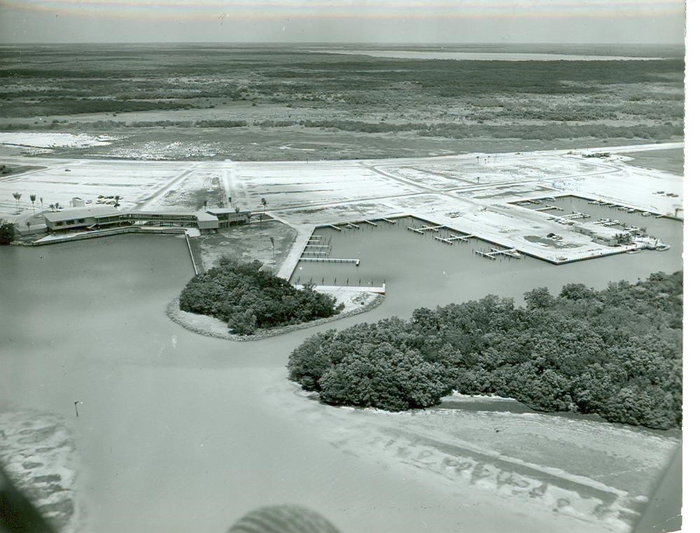 black and white aerial photo of a large parking lot with boat docks and a marina carved into the adjacent water. Mangroves thickets are found near the development.