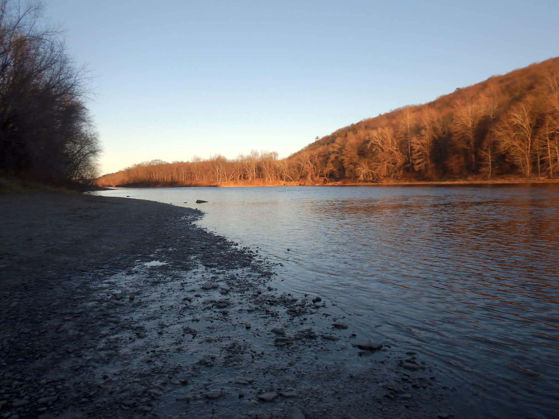River and hillsides covered in trees. River is in shadow and flows toward lower right. Low sun lights trees on far bank in orange glow. Trees are bare of leaves due to the late fall season.