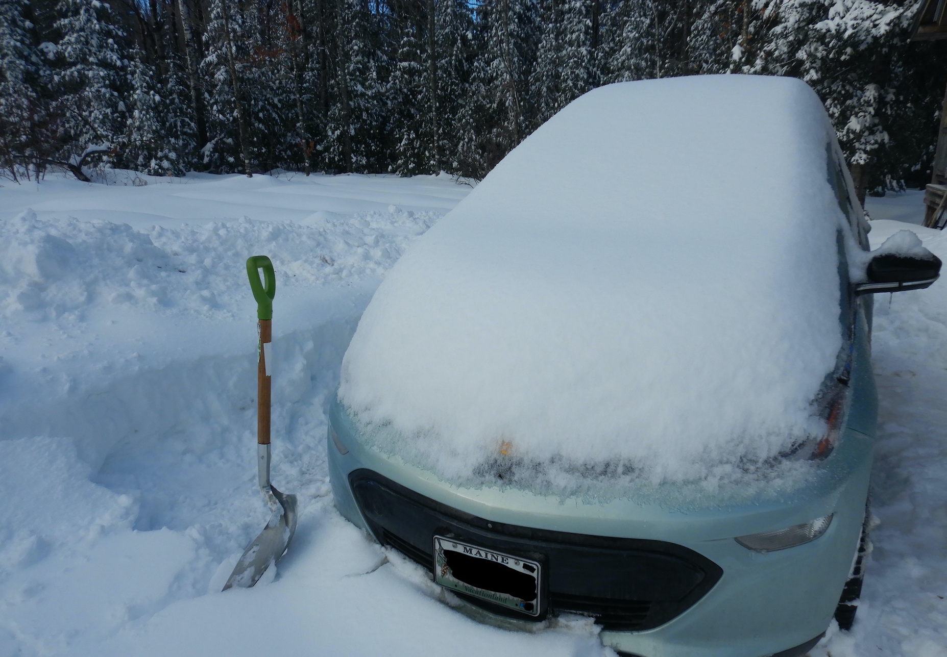 Car parked in deep snow. Car remains snow covered. A snow shovel sticks upright in snow to left of car. A row of snow-covered trees fill the background.