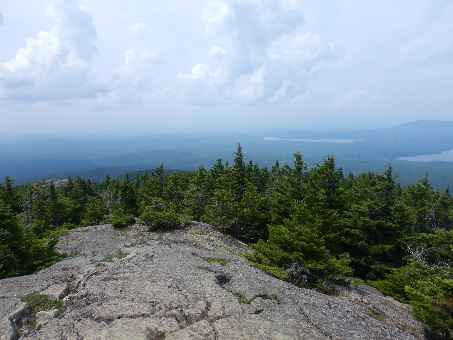 View of forested landscape from mountain summit. Bare rock covers the ground at bottom while spruce and fir trees slope off the mountain. The lowlands and horizon are obscured by haze in the air.