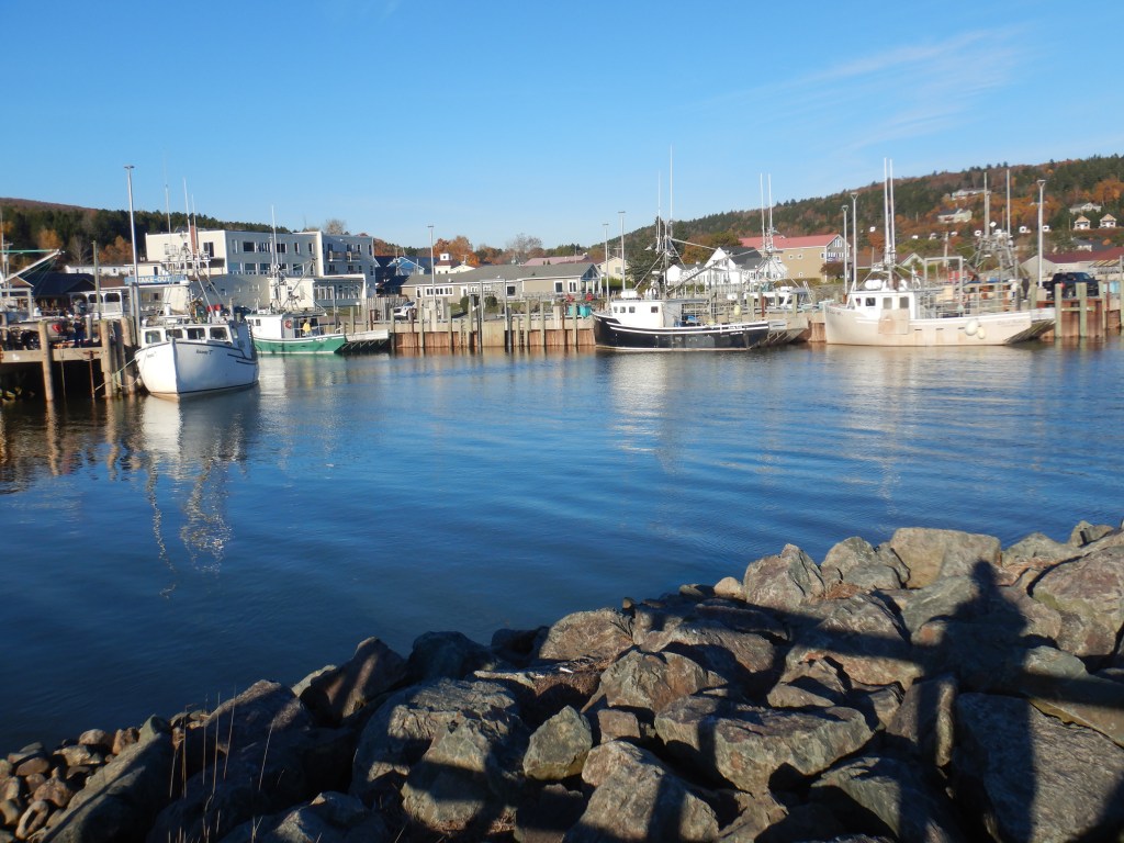 boats float against a pier at high tide. A boulder field of riprap fills the foreground while a small river flows in between.