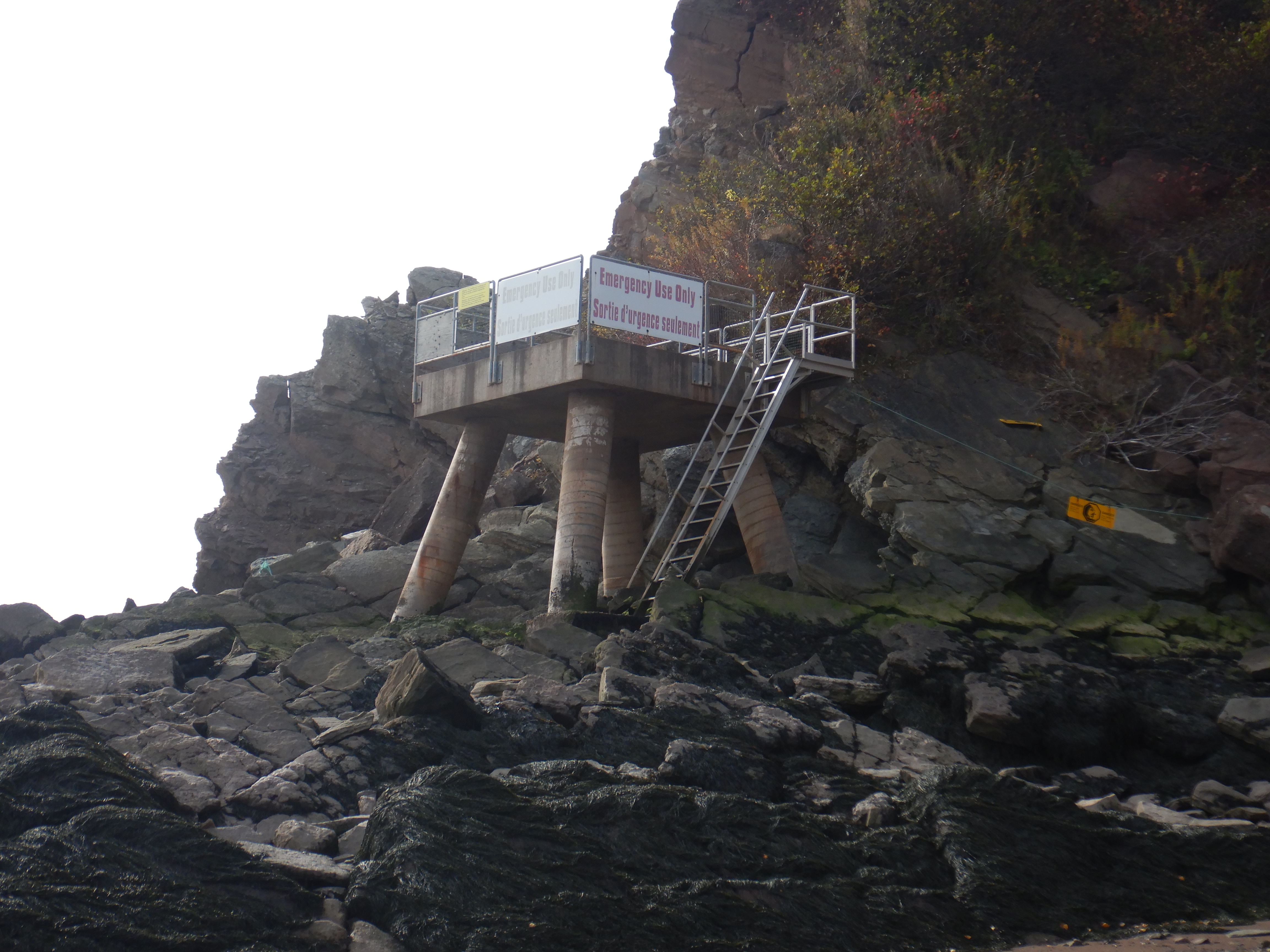 A concrete platform standing on large cyclindrical concrete legs sits against a cliff. A sign on it in red letters says, "Emergency Use Only," and this being Canada, "Sortie d'urgence seulement."